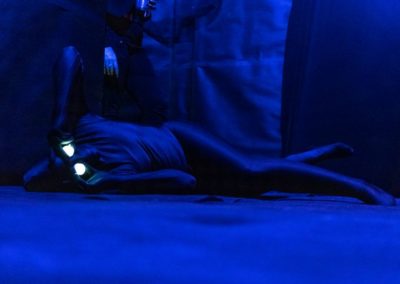 Screen capture from film, Sediment Isotopes. A dark image with blue lighting of a figure laying on the ground in a black body suite holding glowing lights over their eyes.