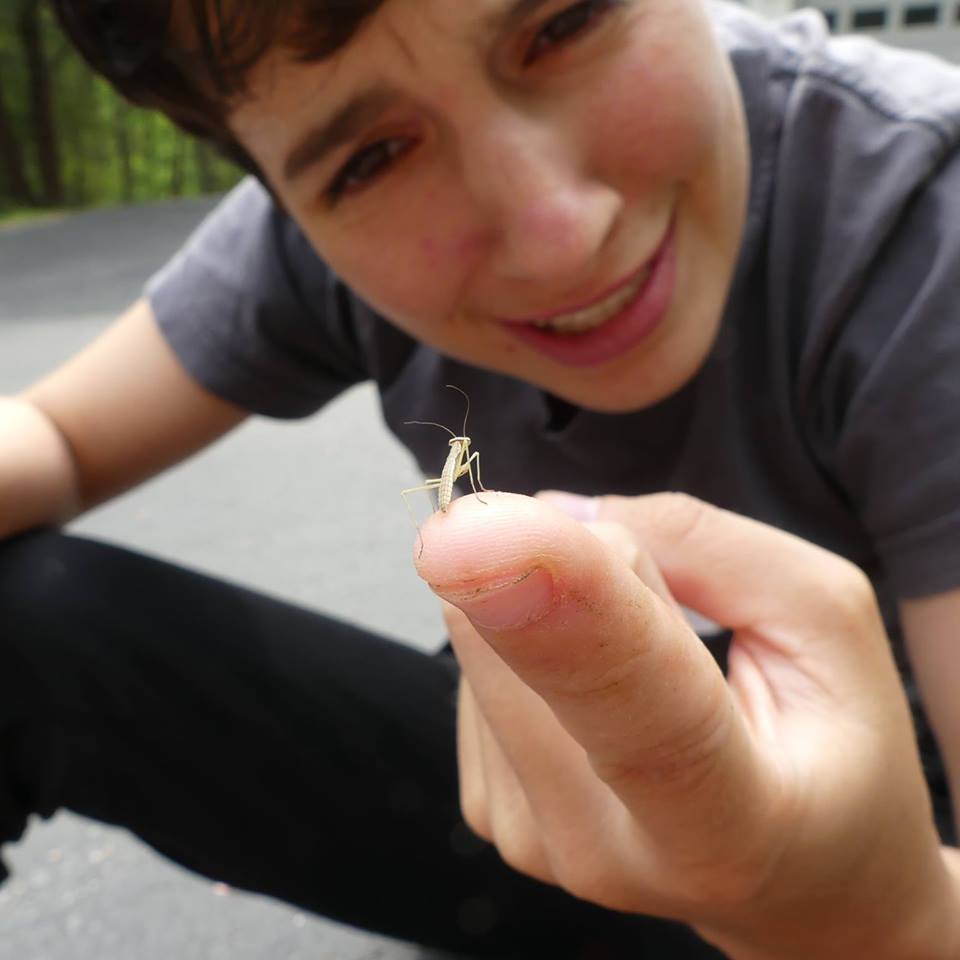 A photo of Olly Silinski outdoors, crouched with a tiny insect on their index finger.