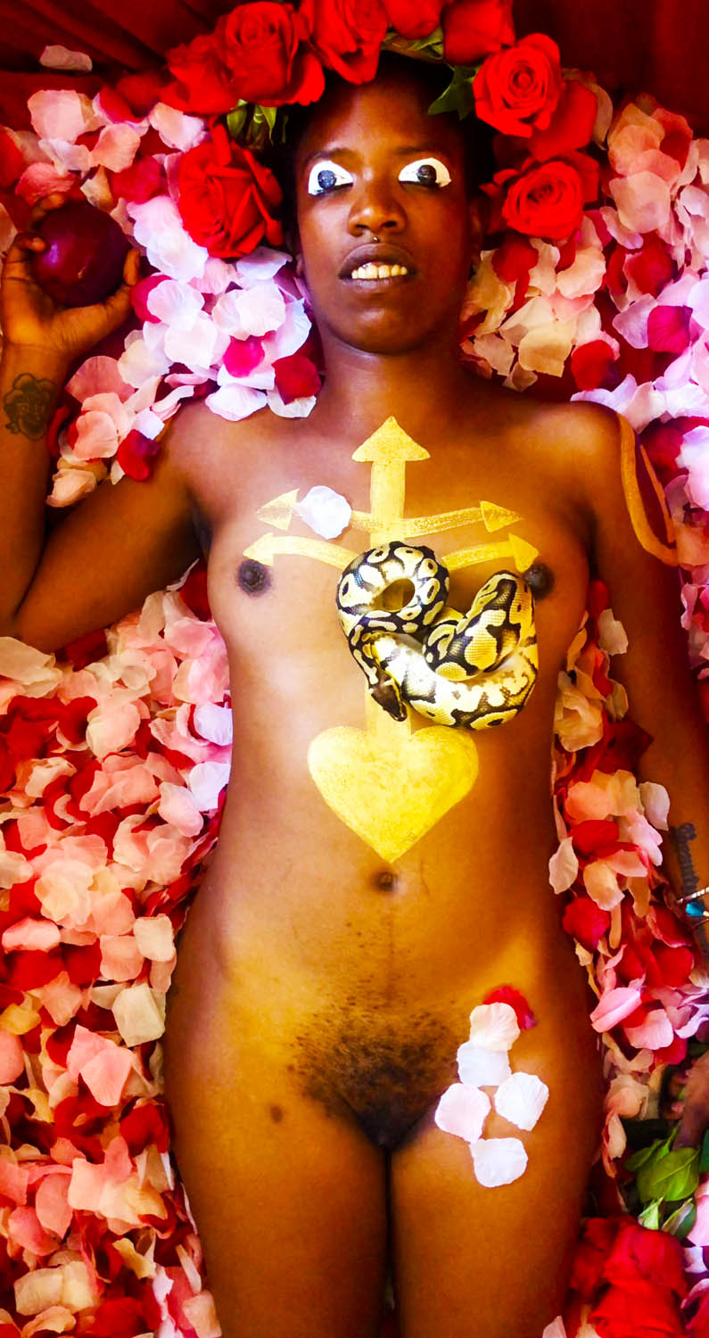 A photo of a nude person laying in a bed of rose petals. They are holding an apple in one hand, have a snake laying on their chest, a crown of flowers on their head, and painted markings on their chest and eye lids.
