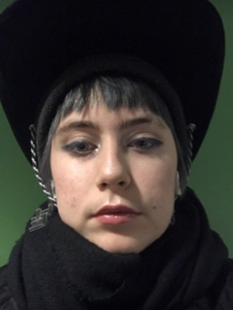 A photo of Clark Woods in front of a green wall. They have short silver hair, and they are wearing a large black hat and a black scarf.