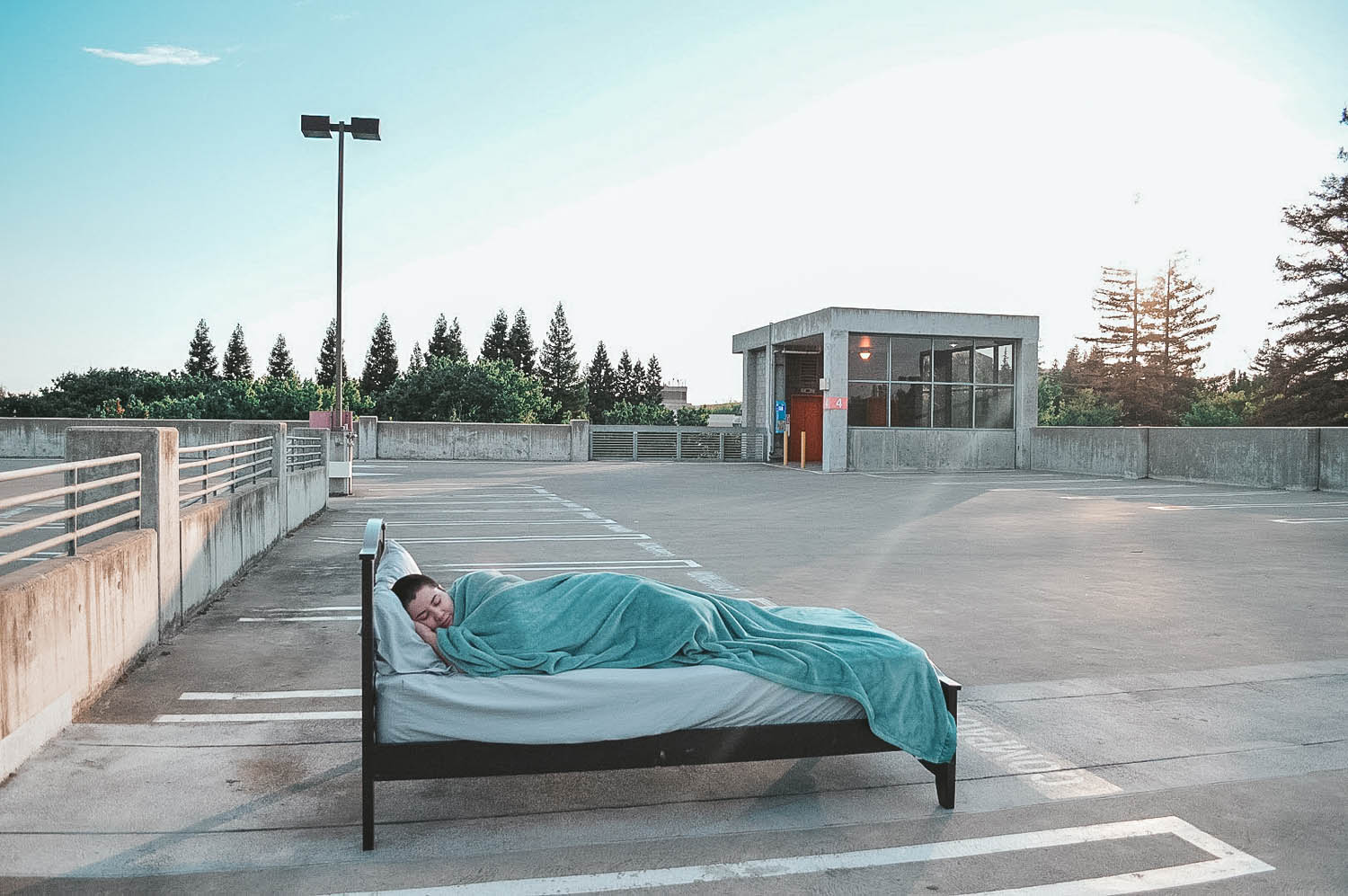 A close up photo of a person sleeping in a bed which is sitting in the middle of an empty parking lot.