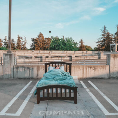 A close up photo of a bed in an empty parking lot. There is a person sleeping in the bed. The photo is looking at the foot of the bed.
