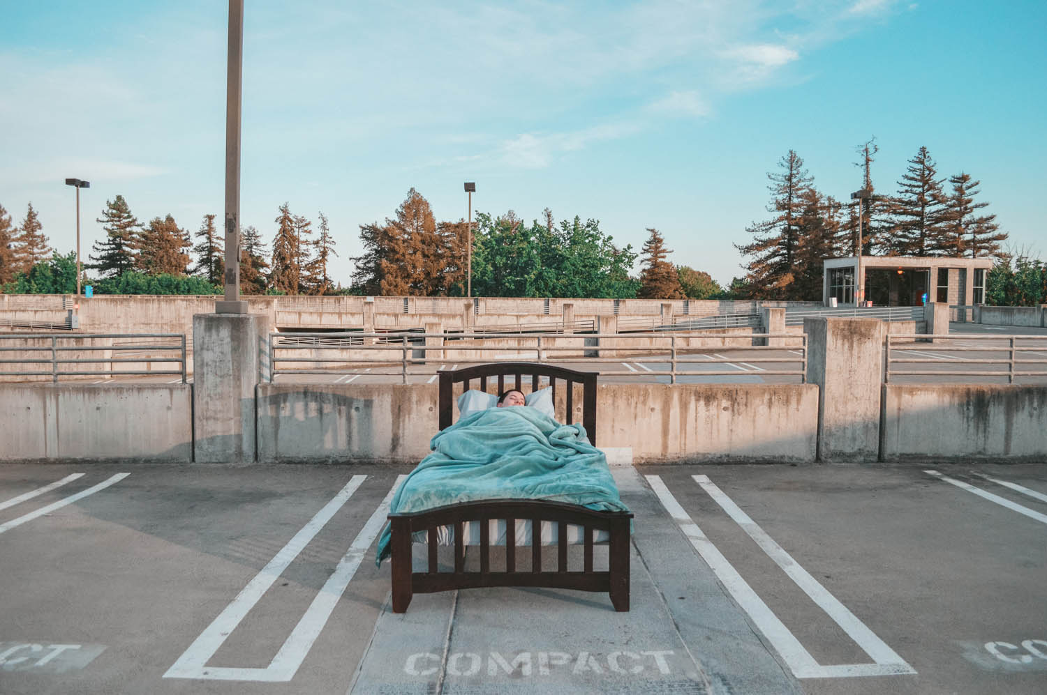 A close up photo of a bed in an empty parking lot. There is a person sleeping in the bed. The photo is looking at the foot of the bed.