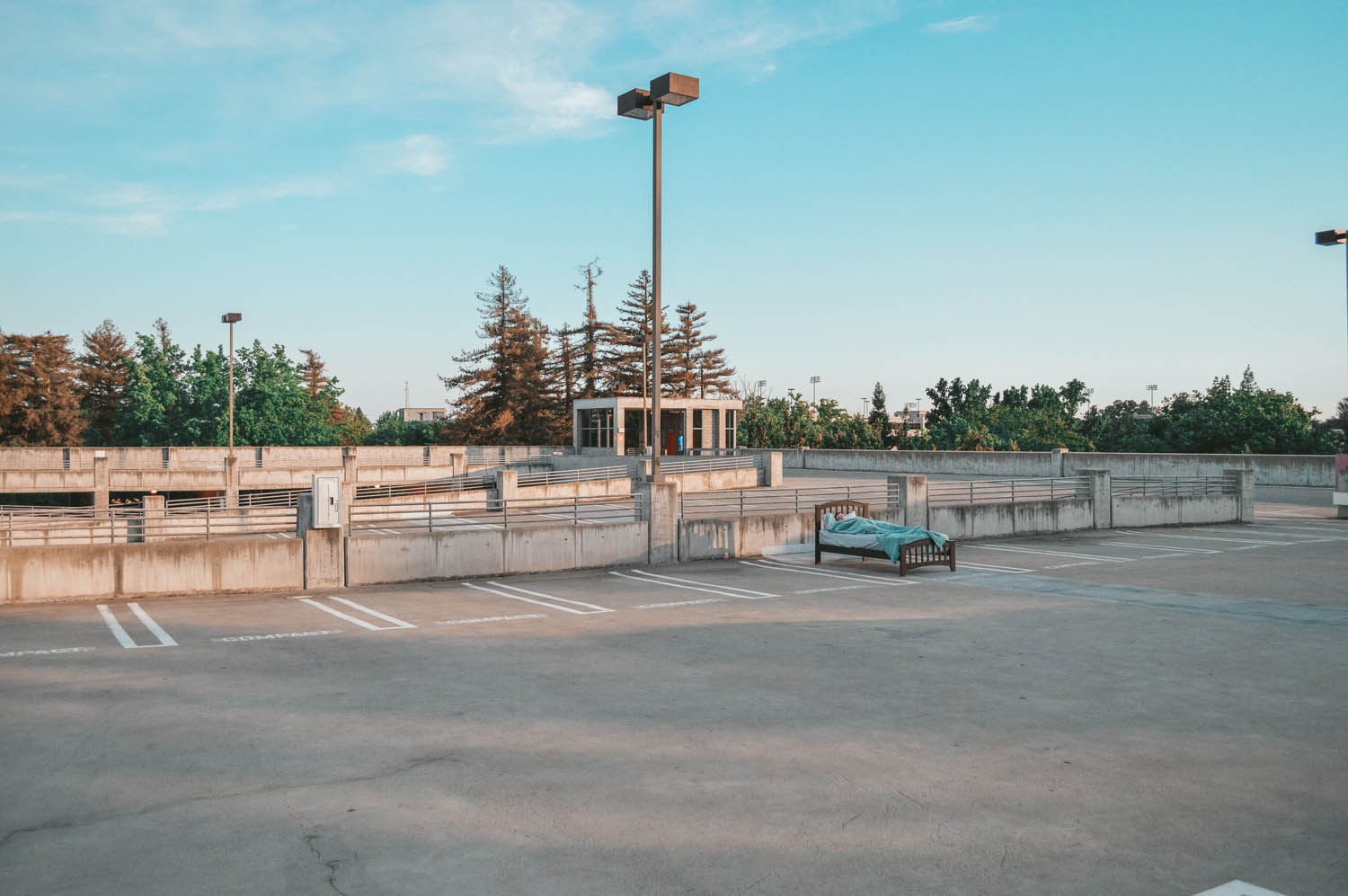 A wide shot photo of an empty parking lot. In the distance, there is a bed in one of the parking spaces with a person sleeping in it.