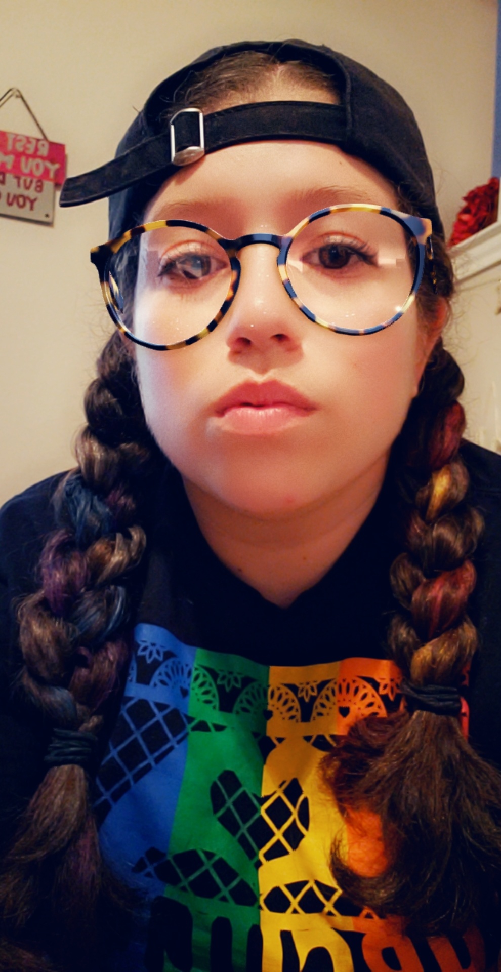 A photo of Sueitko with with snapchat filter glasses, braided hair, backwards hat, and rainbow sweater.