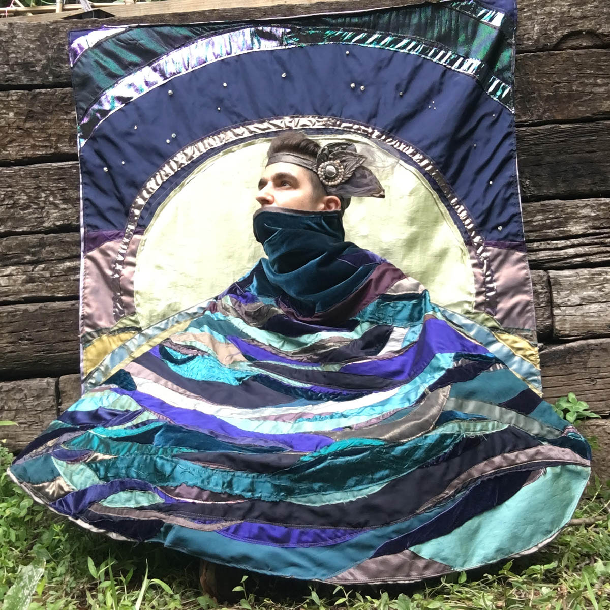 A photo of artist, Eliot eLm Moonstone, posing with their art draped in a multicolored fabric wrap that matches the piece behind them.